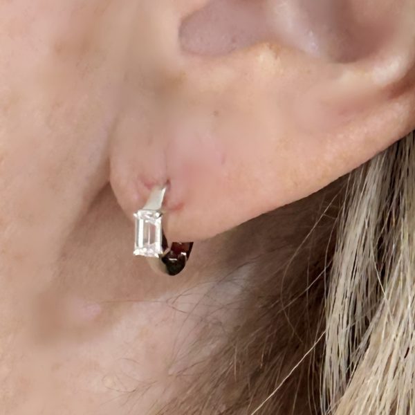 A pair of sterling silver rhodium-plated Huggie earrings featuring a 5x3mm emerald-cut lab-created CZ stone, designed for a sleek and elegant look.