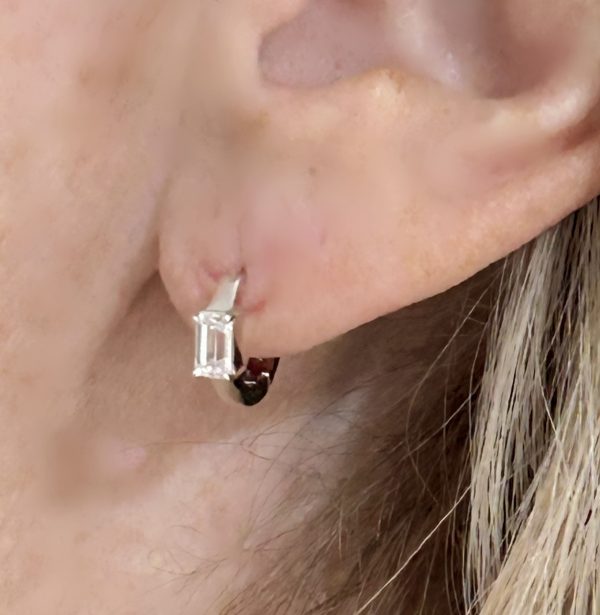 A pair of sterling silver rhodium-plated Huggie earrings featuring a 5x3mm emerald-cut lab-created CZ stone, designed for a sleek and elegant look.